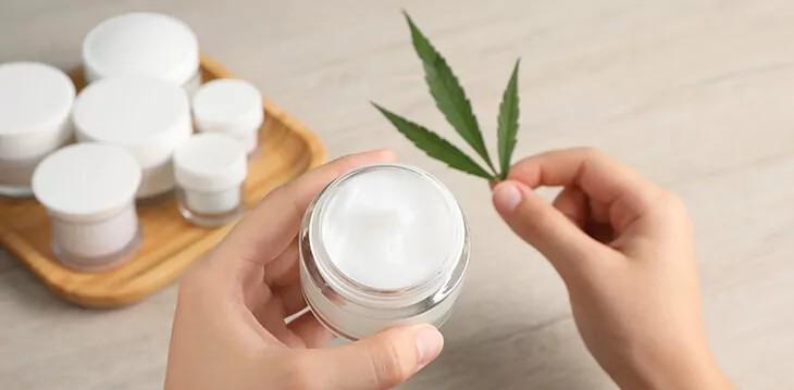 History 6 Femeie ținând în mâini un borcan cu cremă albă hidratantă cu extract CBD.