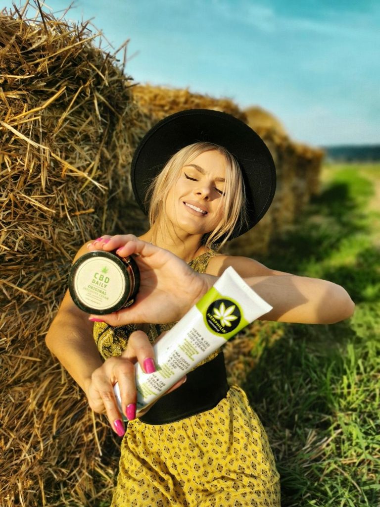 Femeie zâmbind în natură și arătând crema intensivă cu extract CBD pentru piele.