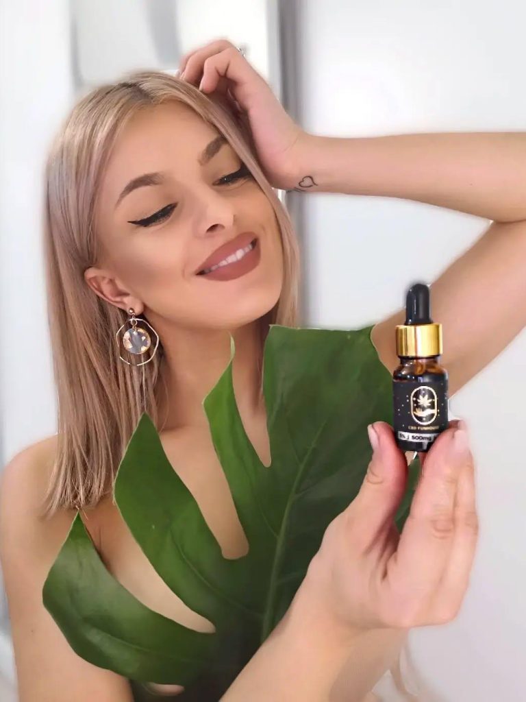 Femeie pozând elegant cu o sticluță de ulei CBD și o frunză mare verde decorativă.